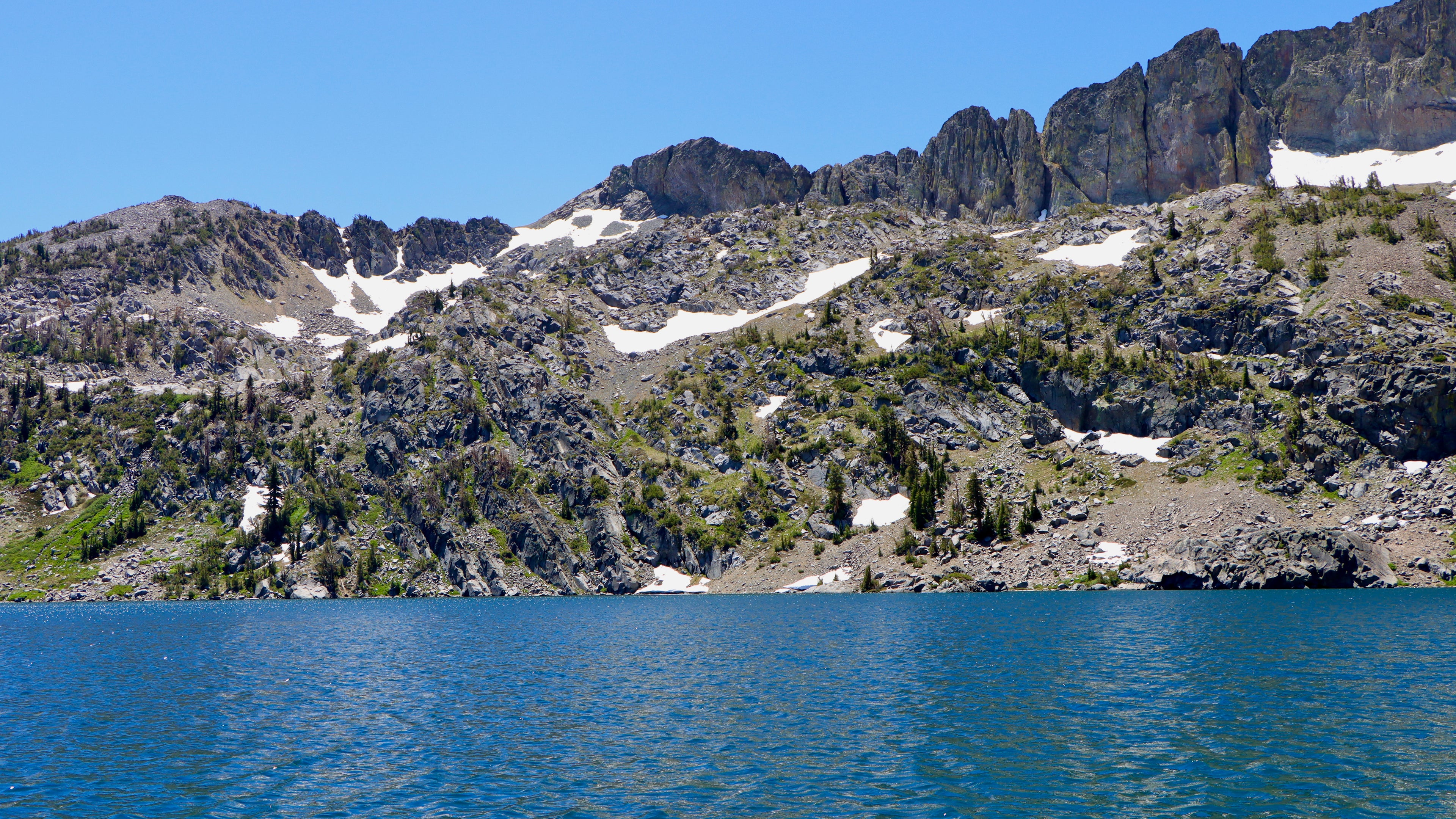 Lake with mountainous landscape and scattered snow patches under a clear blue sky.